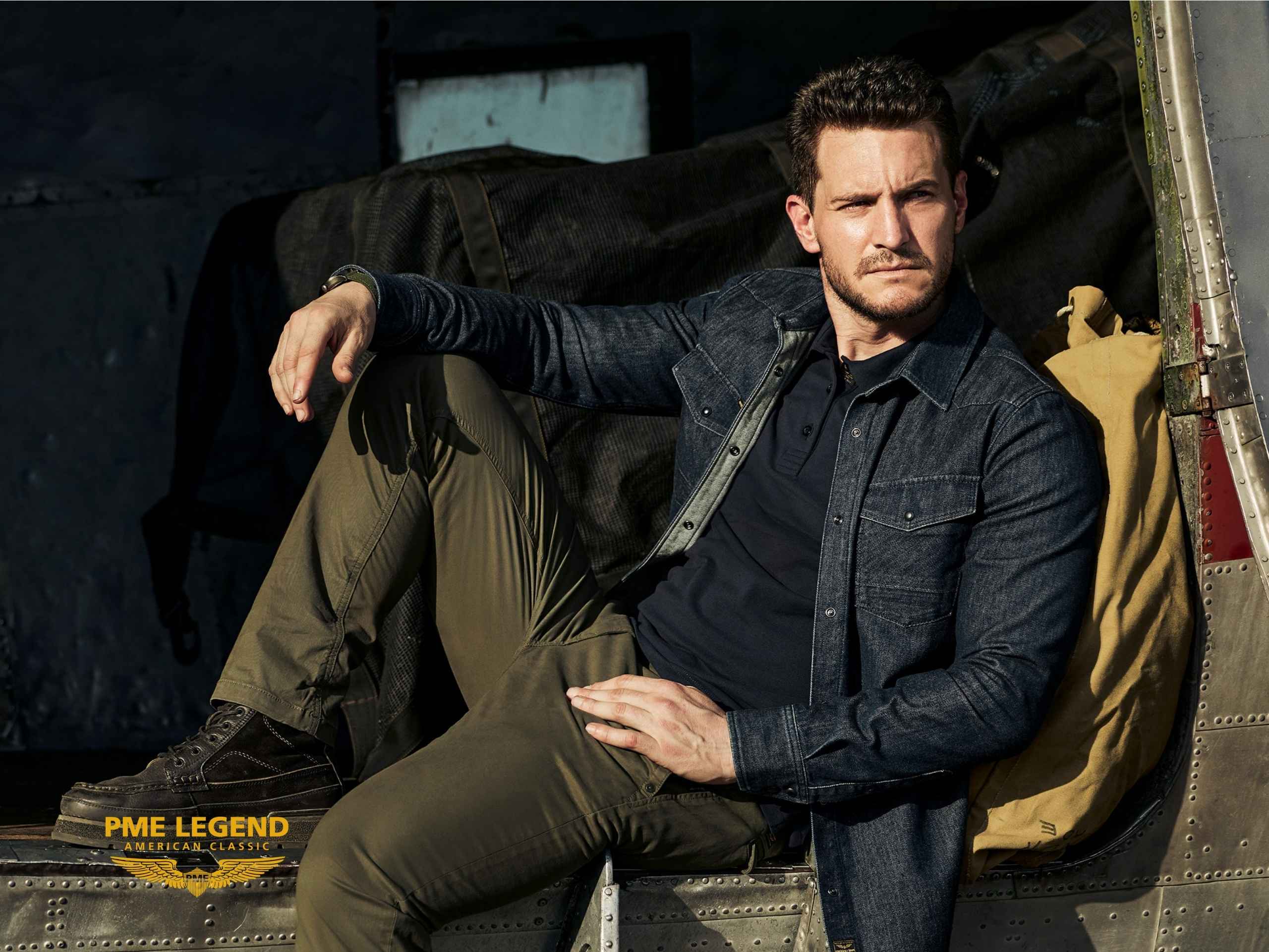 Ein Mann in einem Jeanshemd und einer Khakihose sitzt entspannt im hinteren Teil eines Flugzeugs. In der Ecke ist das Logo „PMI LEGEND AMERICAN CLASSIC“ zu sehen. Die Beleuchtung ist hell und effektvoll.