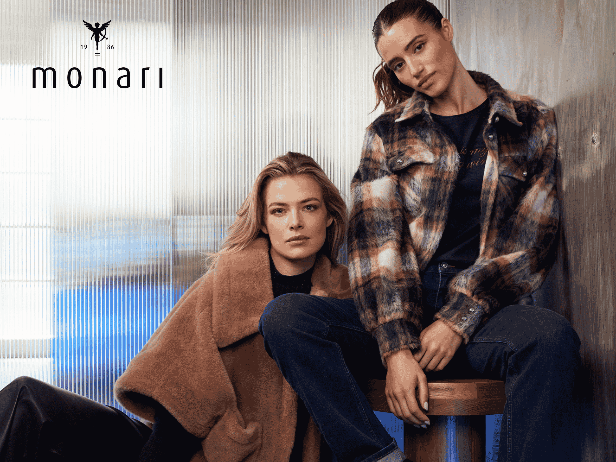 Zwei Frauen posieren in einem Innenraum in stilvollen Herbstoutfits; die eine trägt einen hellbraunen Kunstpelzmantel, die andere eine karierte Jacke über einem schwarzen Shirt. In der oberen linken Ecke ist das Logo der Marke „monari“ zu sehen.